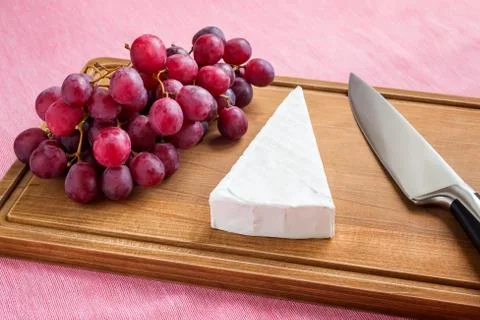 Triangular piece of Brie cheese, red sweet grapes and a chef knife. Stock Photos