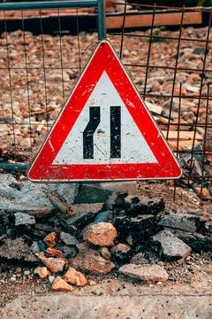 Triangular road narrowing traffic warning sign sitting amidst broken asphal.. Stock Photos