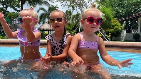 Triplet sisters sitting in a swimming pool, behind a palm tree, wearing 库存影片 266001253