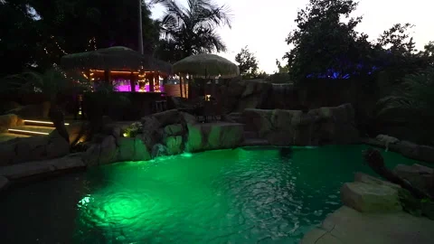 Tropical backyard pool with tiki bar and rock waterfall at dusk Stock Footage 345537183