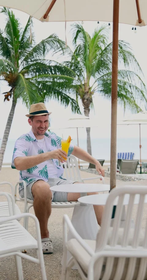 Tropical beach scene with man in hat holding cocktail, sitting under Stock Footage 314044211