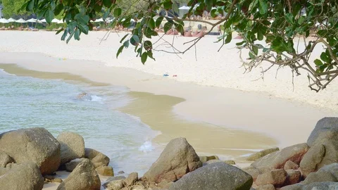 Tropical beach with small waves and sandals in sand behind rock and trees Stock Footage 114904675