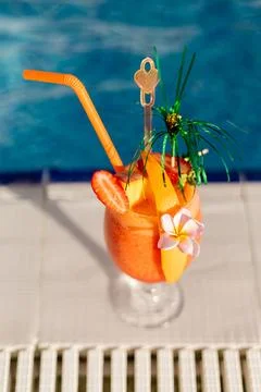A tropical drink is placed on a table beside a pool. The drink has orange and Stock Photos