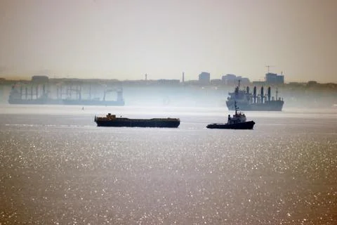 A tug pulls a barge against the backdrop of the city and ships at sea Photos