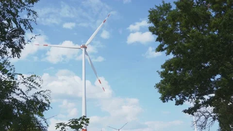 Turning wind turbine seen through lush trees with clouds in blue sky 스톡 동영상 203917491