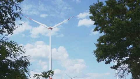 Turning wind turbine seen through lush trees with clouds in blue sky 스톡 동영상 203917508