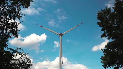Turning wind turbine seen through lush trees against cloudy blue sky Stock Footage 204037399