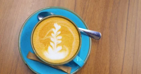 Turquoise ceramic coffee cup, filled with creamy latte art coffee, sits on a Stock Footage 314467765