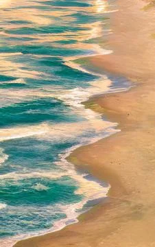 Turquoise ocean waves gently lap on a sandy beach at sunset Stock Photos