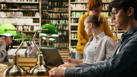 Tutor and student discuss research beside laptop and stacked books in library Stock Footage 336358821