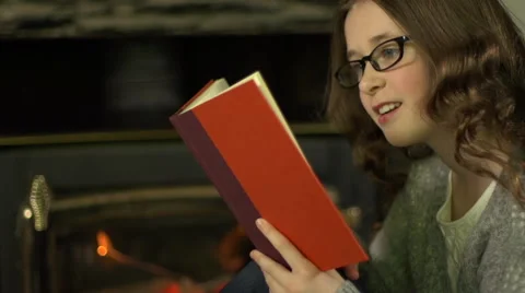 A tween girl sits in front of a fire and reads a book with reading glasses. Stock Footage 44117603