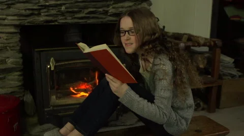 A tween girl sits in front of a fire and reads a book with reading glasses. Stock Footage 44117620