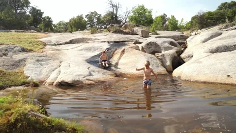 Two 9-year-old Caucasian boys playing in a river by going down a stone slide Stock Footage 277101294