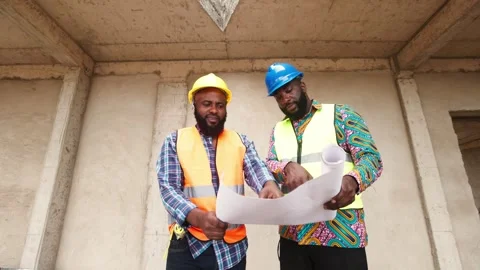 Two African engineers in hard hats, vests review blueprints on site 库存影片 346257680
