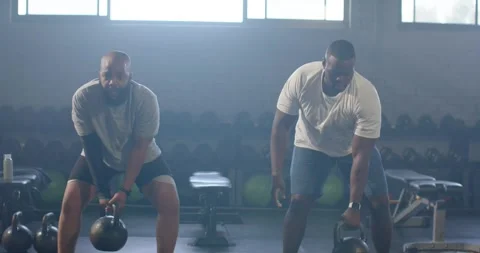 Two African men in tees doing kettlebell swings starting from holding bells for Stock Footage 342493929