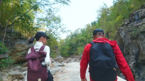Two asian hiker stands on a light colored rock beside a gentle flowing stream Видео 342409257