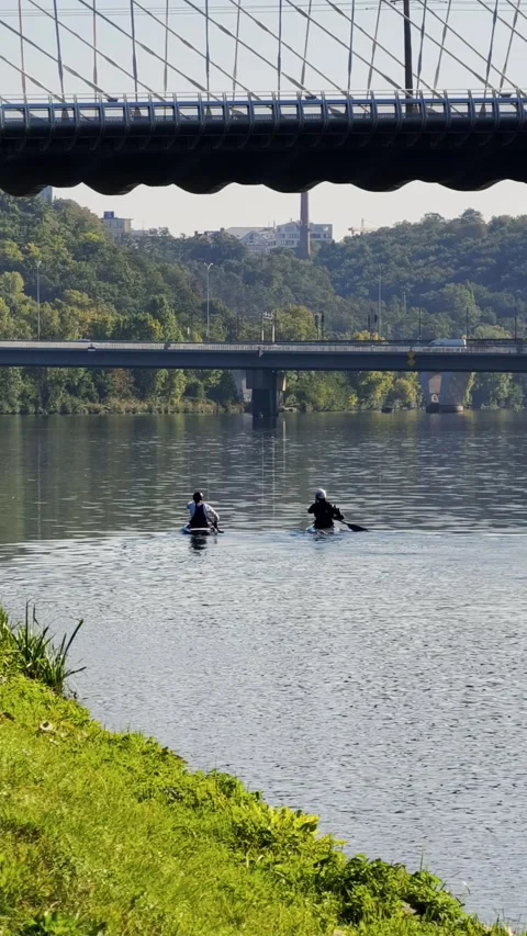 Two athletes kayaking across a calm river with bridges in the background Stock Footage 346612156