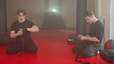 Two athletes kneel on red mat in gym wrapping hands with black straps Stock Footage 322551961
