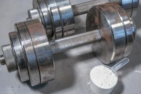 Two athletic iron dumbbells lie on a concrete floor next to a spoon of protei Foto stock