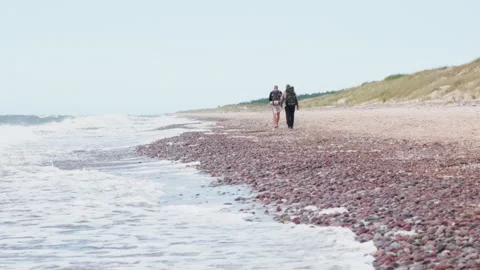 Two backpackers walking along pebble beach by the sea Stock Footage 346525384