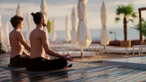 Two bare-chested guys in black shorts sit and meditate on a private beach in the Stock Footage 249234758
