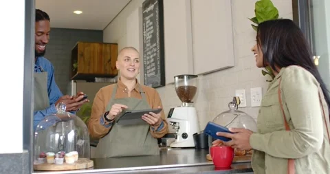 Two baristas serving Indian customer at counter, placing red mug, tapping card Stock Footage 337537249