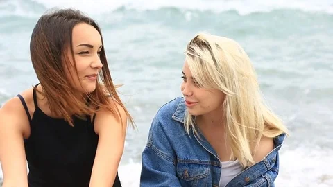 Two beautiful girlfriends sitting on the beach against the waves of the sea and Stock Footage 77030486