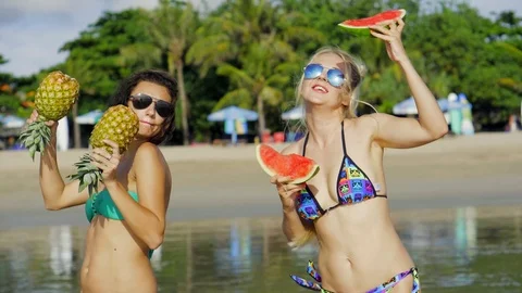 Two beautiful sexy daring lesbians, sunglasses keep tropical fruit on the beach Stock Footage