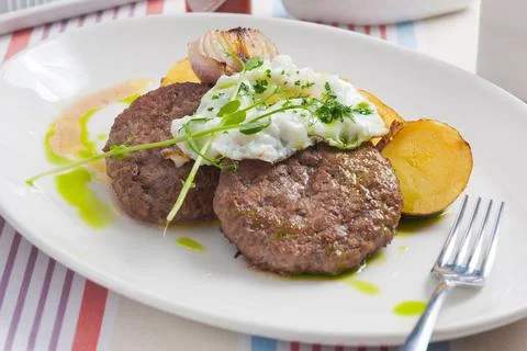 Two beef steaks with potatoes, fried egg and baked onions on a white plate Stock Photos