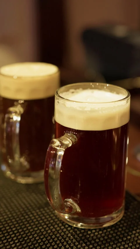 Two beer mugs with dark ale and creamy foam on bar counter under warm pub Stock Footage 345270432