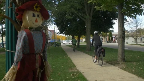 Two Bicyclists Passing By Friendly Scarecrow On A Sunny Autumn Day, 60fps Stock Footage 124620404