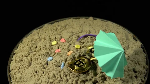 Two bitcoins in the sand under a small umbrella and rotate on a black background Stock Footage 88886814