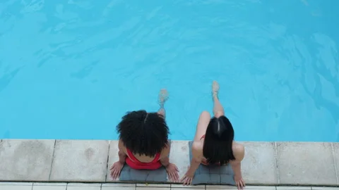 Two black haired girlfriends swing legs in water in public pool relax smile look Stock Footage 137022455
