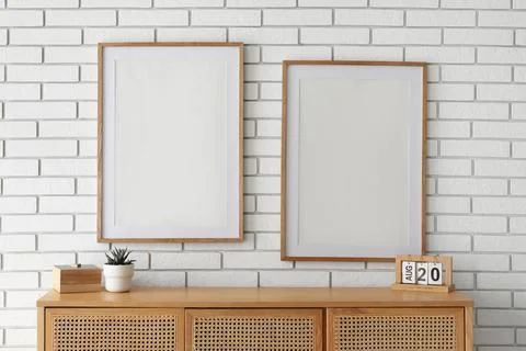 Two blank wooden frames on a white brick wall above a wooden cabinet with a p Stock Photos