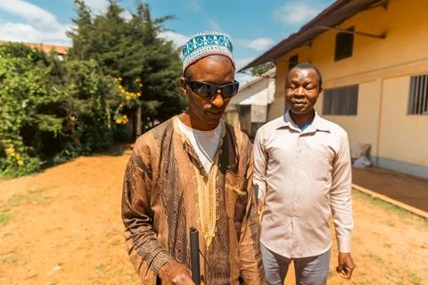 Two blind men standing in front of a house, one wearing a turban and sunglass Stock Photos