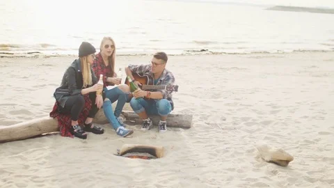 Two blondes and a guy drinking alcohol by the fire sitting on a log while sunset Stock Footage 75681431