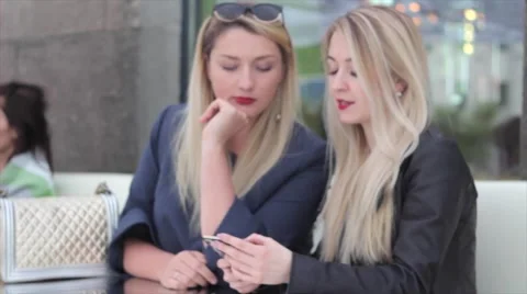 Two blondes are sitting in a café and discuss photos in the smartphone Vídeos de archivo 64231488