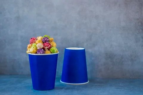 Two blue paper cups with multi-colored caramel popcorn on gray background. Stock Photos