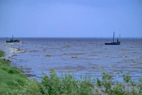 Two boats float on calm, wide water. Green grass lines the shoreline. Blue sk Stock Photos