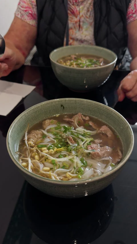 Two bowls of beef noodle soup on restaurant table with bean sprouts, herbs, and Stock Footage 345453032