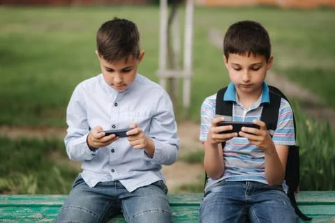 Two boy sitting on the bench and play online games. One boys with backpack Stock Photos