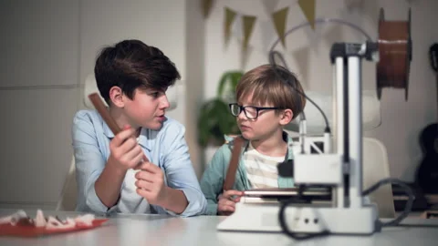 Two boys are sitting at a table with a working 3D printer in the foreground Stock Footage 335373291
