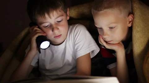 Two boys at night  under a blanket reading a book with a flashlight Stock Footage 56184906