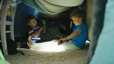 Two boys play with flashlights and walkie talkies inside homemade blanket fort Stock Footage 127549737