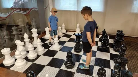 Two boys playing chess on large life-sized chessboard surrounded by big black Stock Photos