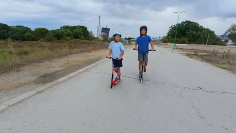 Two boys riding scooters on an empty road outdoors, wearing helmets, active Stock Footage 318160274