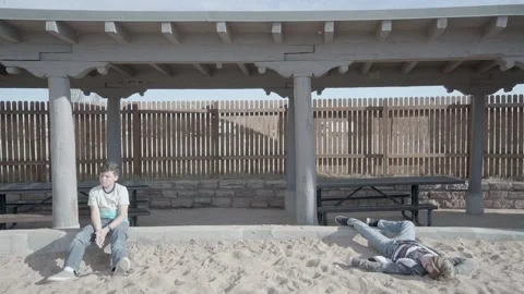 Two boys sit in sand under fenced patio pavillion area playing hooky. Video stock 169734412