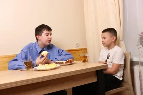 Two boys at table, one in blue shirt eating bread, other listening with fork  Stock Photos