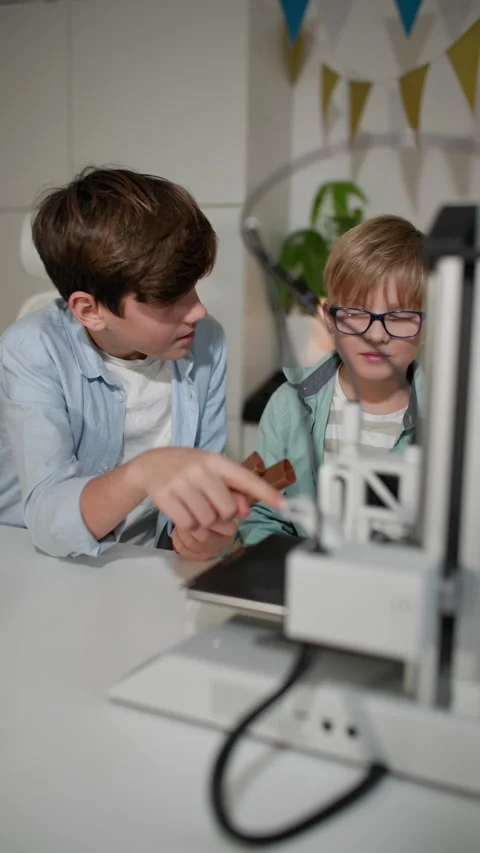 Two boys at a white table chat, and watch a 3D printer in operation Stock Footage 335390074