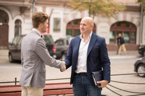Two businessmen shaking hands while standing in front of a classic European b Stock Photos